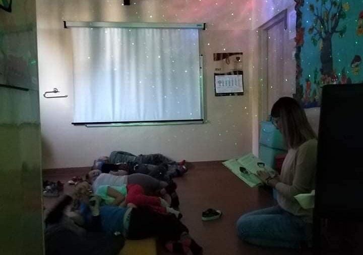 Na zdjęciu dzieci leżące na materacach, obok pani nauczycielka czyta dzieciom książkę. Na suficie wyświetlone gwiazdy
