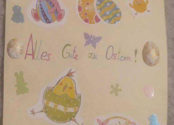Kartka wielkanocna. Na środku kartki napis w języku niemieckim Wszystkiego najlepszego z okazji Świąt Wielkanocnych. Na całej kartce przyklejone naklejki - pisanki, kurczak, baranek