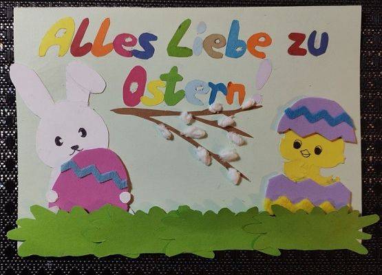 Kartka wielkanocna. Na górze kartki napis w języku niemieckim Wszystkiego najlepszego z okazji Świąt Wielkanocnych. Po prawej stronie kartki królik trzyma pisankę, po prawej stronie kurczaczek w jajku, na środku gałązki bazi