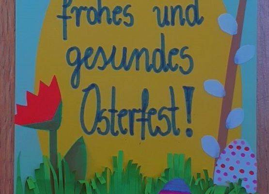Kartka wielkanocna. Na niebieskiej kartce żółte jajko a na nim napis w języku niemieckim szczęśliwych i zdrowych Świąt Wielkanocnych. Po lewej stronie jajka czerwony kwiatek, po prawej gałązka bazi. Na dole trawa z papieru, w której ukryte są