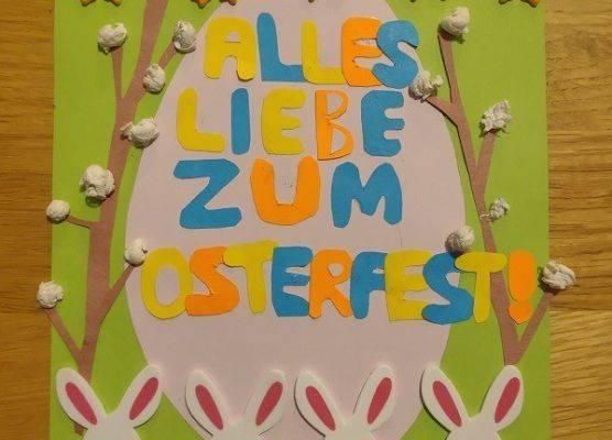 Na środku zielonej kartki naklejone różowe papierowe jajko. Na jajku napis w języku niemieckim Wszystkiego najlepszego z okazji świąt Wielkanocnych.Po obu stronach jajka wycięte z papieru bazie. Na górze kartki 5 kurczaczków, na dole kartki 4
