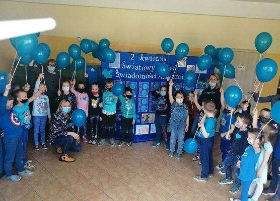 Dzieci z oddziałów przedszkolnych ubrani na niebiesko. W ręku trzymają niebieskie balony. W tle na niebieskim materiale biały napis - 2 kwietnia Światowy Dzień Świadomości Autyzmu