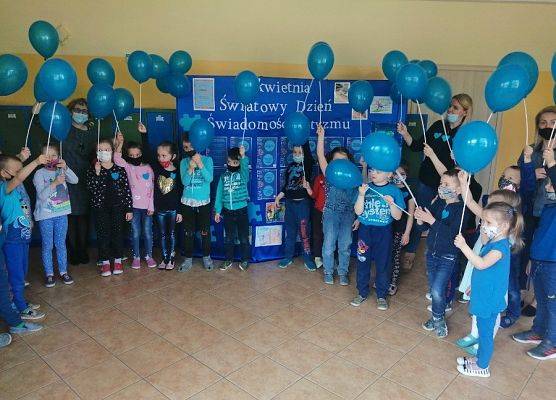 Dzieci z oddziałów przedszkolnych wraz z wychowawcami, ubrani na niebiesko. W ręku trzymają niebieskie balony. W tle na niebieskim materiale biały napis - 2 kwietnia Światowy Dzień Świadomości Autyzmu