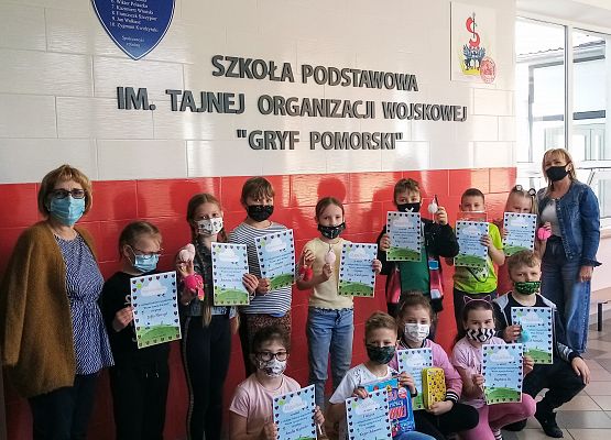 Laureaci szkolnego konkursu recytatorskiego dla klas 1-3 pod tytułem  "Wiosna w poezji dziecięcej". Uczniowie trzymają dyplomy i nagrody. Z prawej strony stoi pani dyrektor, z lewiej nauczycielka. W tle białoczerwona tablica pamiątkowa