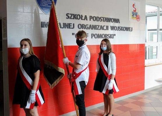 Poczet Sztandarowy Szkoły na tle biało czerwonej ściany pamiątkowej.