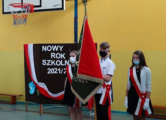 Rozpoczęcie roku szkolnego na sali gimnastycznej. Poczet sztandarowy szkoły w tle brązowy parawan z napisem Nowy Rok Szkolny 2020 - 2021 udekorowany flagą Polski