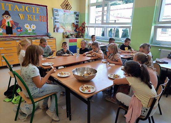 Uczniowie siedzą przy stołach i jedzą sałatkę owocową