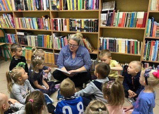 Dzieci z oddziału 5 -latków w bibliotece szkolnej. Dzieci siedzą blisko bibliotekarki, która czyta im książkę. Wtle regały z książkami