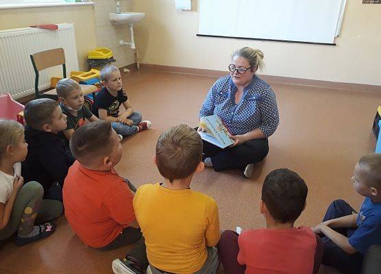 Bibliotekarka w sali lekcyjnej 6-latków. Dzieci siedzą blisko bibliotekarki, która czyta im książkę.