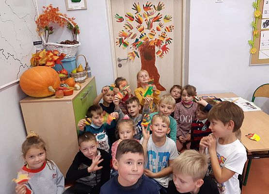 Na zdjęciu grupa dzieci siedzących na podłodze. W tle na drzwiach wejściowych do klasy przyklejone jest jesienne drzewo, którego liście zrobione są odbitych dłoni uczniów