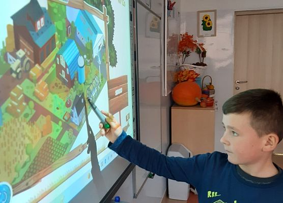 Uczeń klasy 1 stoi przed tablica multimedialną, w ręku trzyma pisak interaktywny i wskazuje na wyświetlany na tablicy rysunek gospodarstwa rolnego.