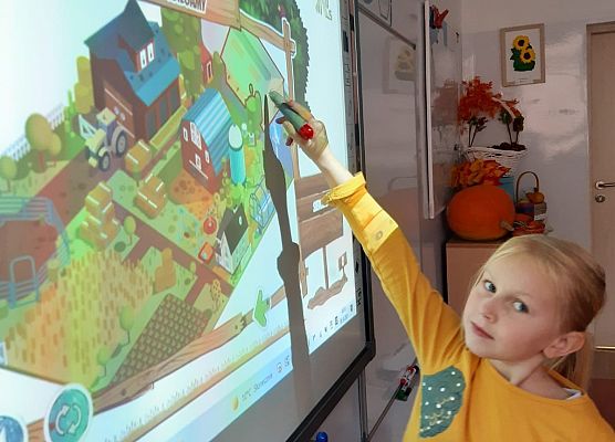 Uczennica klasy 1 stoi przed tablica multimedialną, w ręku trzyma pisak interaktywny i wskazuje na wyświetlany na tablicy rysunek gospodarstwa rolnego.