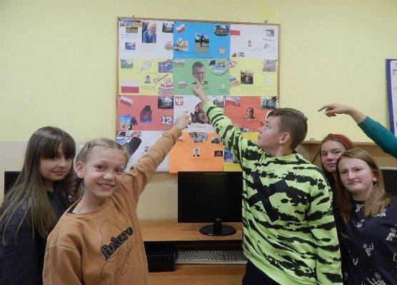 Piecioro uczniów klasy 7 wskazuke na tablicę korkową, na której są prace dotycżące projektu na temat przedstawiania się w języku niemieckim