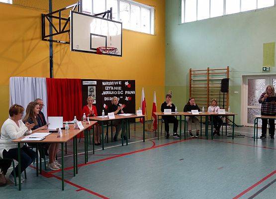 Na sali gimnastycznej ustawione są stoliki przy których siedzi 9 osób. Jedna osoba trzyma mikrofon i czyta. W tle dwa parawany. Jeden przykryty biało-czerwonym płótnem, na drugim przyczepiony napis narodowe