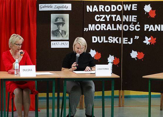 Przy stolikach siedzą dwie kobiety. Obie trzymają mikrofony i czytają. W tle parawany. Jeden przykryty biało-czerwonym płótnem, na drugim biały napis Narodowe czytanie Moralność pani Dulskiej oraz zdjęcie
