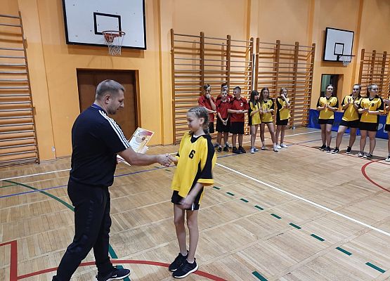 Sala gimnastyczna. Mężczyzna wręcza dyolom zawodniczce. W tle drużyny koszykarskie