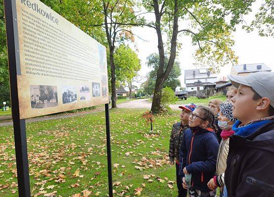 Na świeżym powietrzu, w parku. Uczniowie stoją przodem do tablicy opisującej historię Redkowic. Czytają.