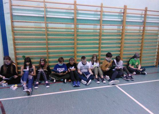 Grupa uczniów siedzi na ziemi w sali gimnastycznej i czytają książki. W tle drabinki gimnastyczne