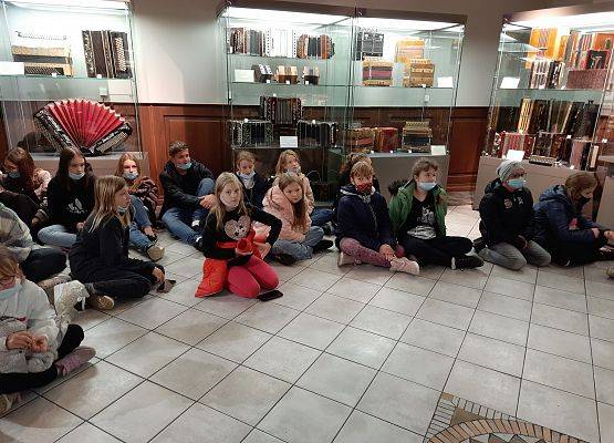 Uczniowie w Muzeum Akordeonu. Dzieci siedzą na podłodze, słuchają prelekcji. W tle eksponaty za szybą