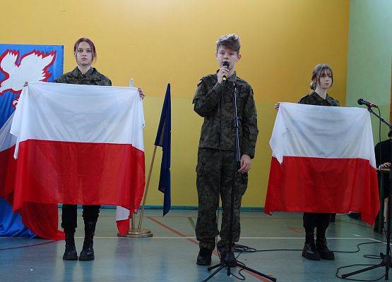 Uczeń w żołnierskim stoju stoi przed mikrofonem. Recytuje wiersz. Po obu stronach chłopca stoją uczennice z dużymi flagami Polski.