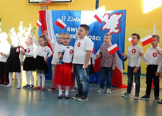 Chłopiec i dziewczynka śpiewają piosenkę. Za nimi stoją dzieci z flagami Polski. W tle dekoracja patriotyczna.