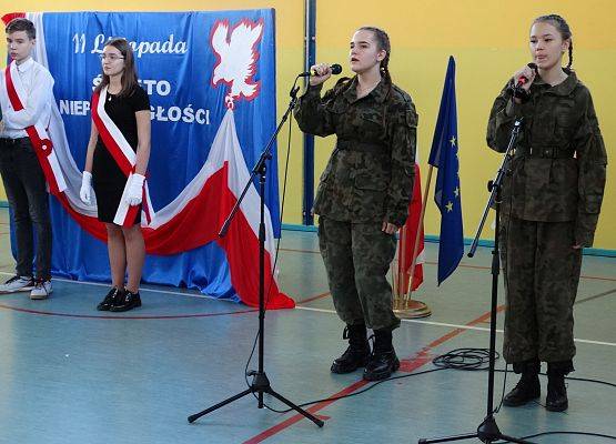 Dwie uczennice w żołnierskich mundurach trzymają w ręku mikrofony. Śpiewają pieśń. W tle dekoracja patriotyczna i poczet sztandarowy szkoły.