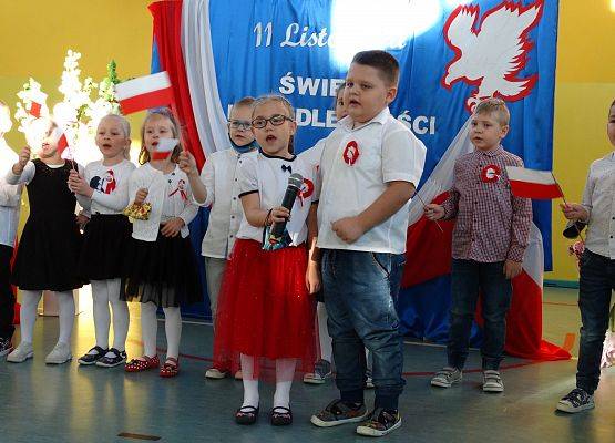 Dziewczynka i chłopiec śpiewają piosenkę. Za nimi stoją dzieci z flagami Polski. W tle dekoracja patriotyczna.