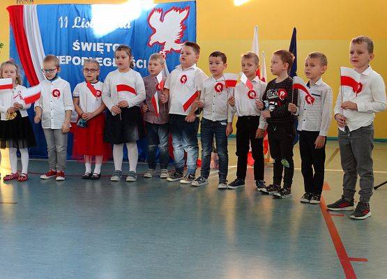 Sala gimnastyczna. Dzieci z oddziałów przedszkolnych recytują wiersz . W tle dekoracja patriotyczna.