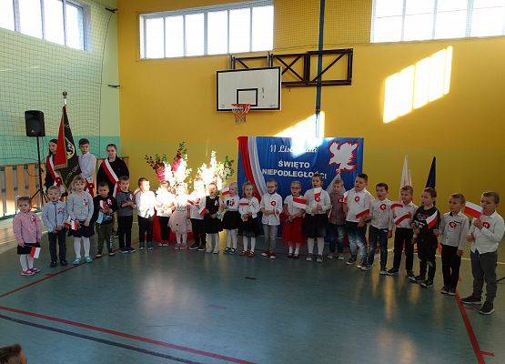 Sala gimnastyczna. Dzieci z oddziałów przedszkolnych stoją w półkolu. Recytują wiersz. W tle dekoracja patriotyczna