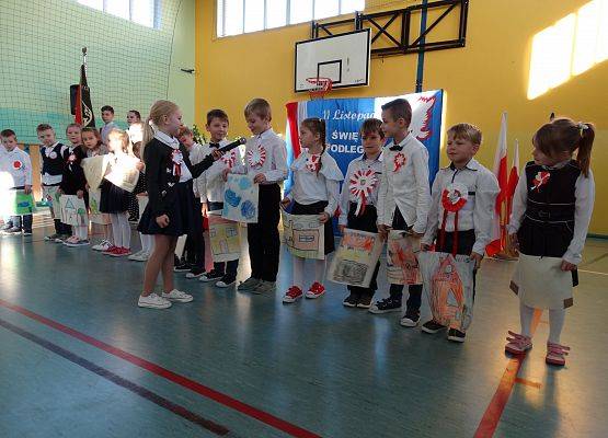 Sala gimnastyczna. Uczniowie klasy pierwszej stoją w jednym rzędzie. Dziewczynka stoi przed nimi i trzyma mikrofon. Dzieci recytują wiersz. W tle dekoracja patriotyczna