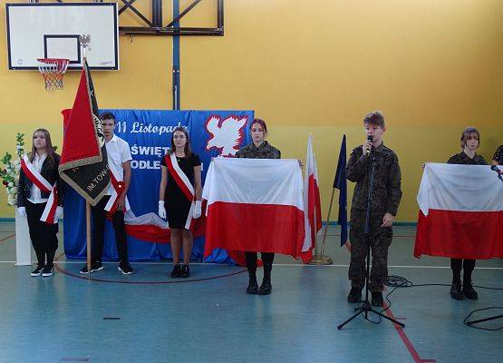 Uczeń w żołnierskim stoju stoi przed mikrofonem. Recytuje wiersz. Po obu stronach chłopca stoją uczennice z dużymi flagami Polski. W tle dekoracja patriotyczna i poczet sztandarowy szkoły.