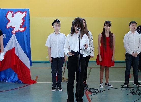 Uczennica stoi przed mikrofonem i czyta wiersz. W tle czwórka uczniów, dwie dziewczynki i dwóch chłopców.