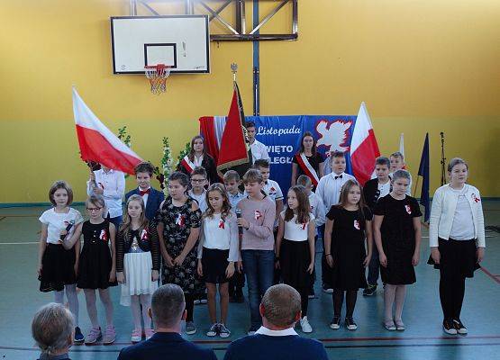 Uczniowie klasy czwartej śpiewają piosenkę. W tle dwie duże flagi Polski.