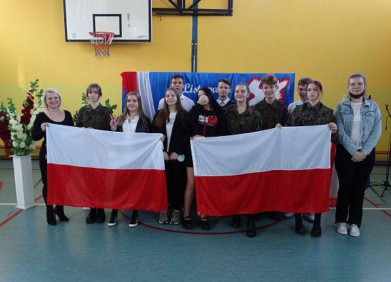 Uczniowie klasy ósmej stoją w grupie wraz z wychowawczynią . Uczniowie z pierwszego rzędu trzymają dwie duże flagi Polski. W tle dekoracja patriotyczna.