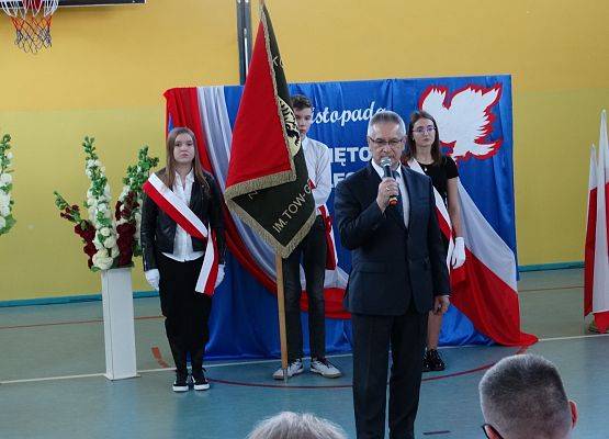 Wójt Gminy Nowa Wieś Lęborska trzyma w dłoni mikrofon i przemawia. W tle dekoracja patriotyczna i poczet sztandarowy szkoły.