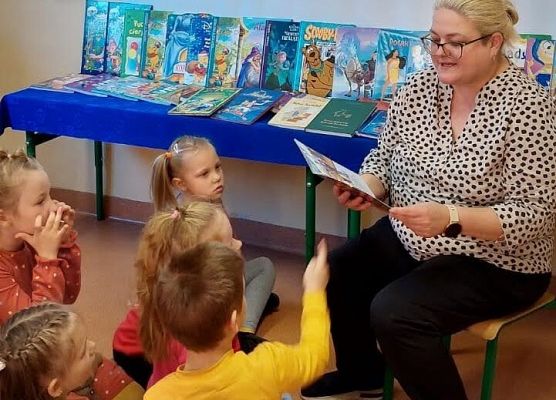 Bibliotekarka czyta przedszkolakom. W tle wystawa książek i plakat akcji Przerwa na czytanie