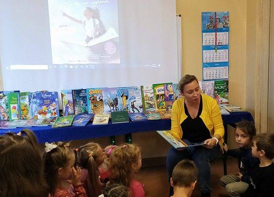 Nauczycielka czyta bajkę przedszkolakom . W tle wystawa książek z bajkami i plakat akcji Przerwa na czytanie