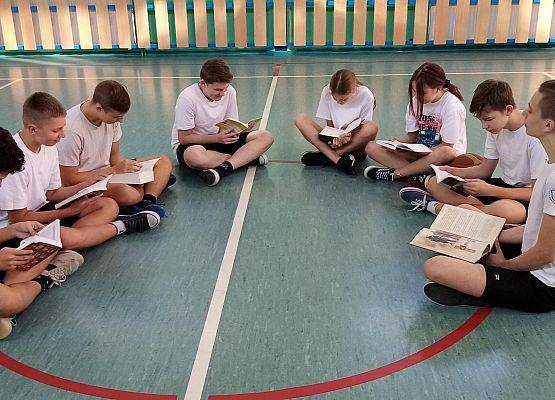 Uczniowie siedzą w kole na sali gimnastycznej i czytają książki.